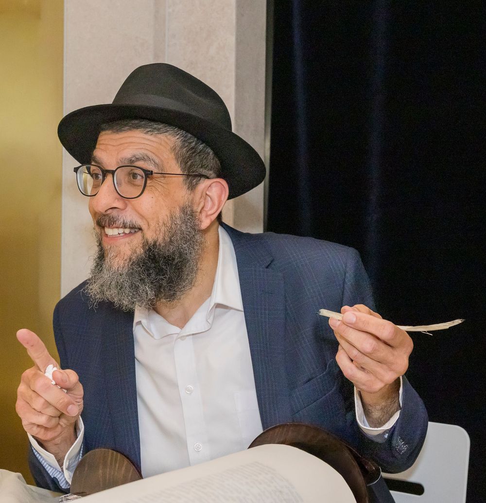 A person in a black hat and navy blazer gestures animatedly while holding utensils during a conversation.