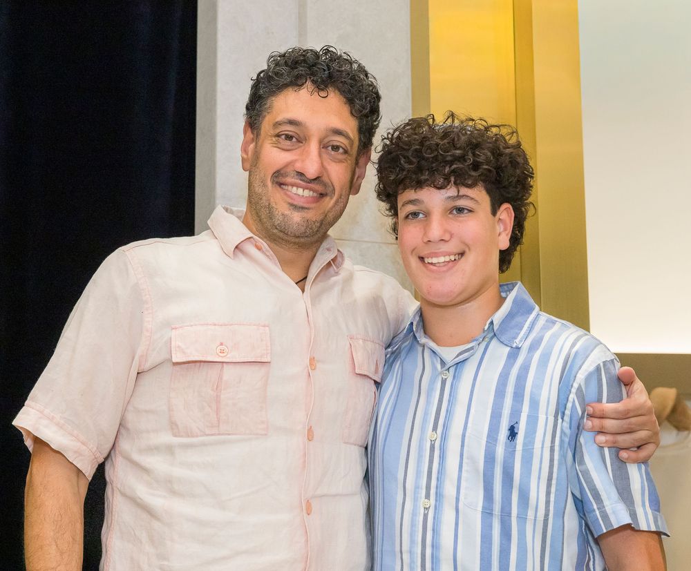 Two people smiling together while standing side by side, wearing button-down shirts in a room with yellow walls.
