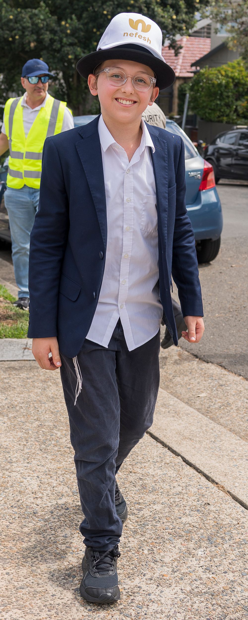 A young student in a navy blazer, white shirt and dark pants walks confidently on a gravel path while wearing a baseball cap.