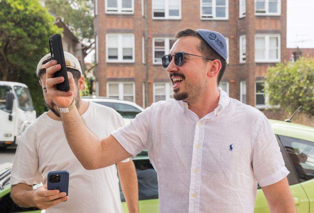 Group of people in white shirts enjoying drinks and socializing outdoors near brick buildings on a sunny day.