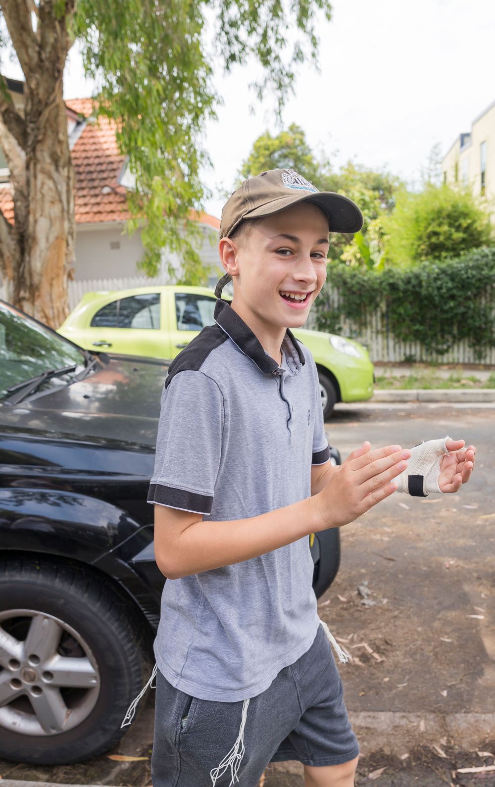 A young person in a grey polo shirt smiles and gestures while standing next to a parked car on a sunny day.