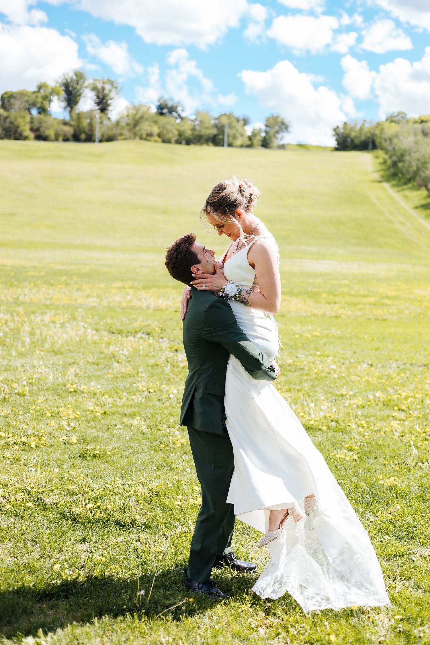 Groom in green suit lifts bride in white wedding dress on sunny green field with blue sky. Destination wedding photographers.