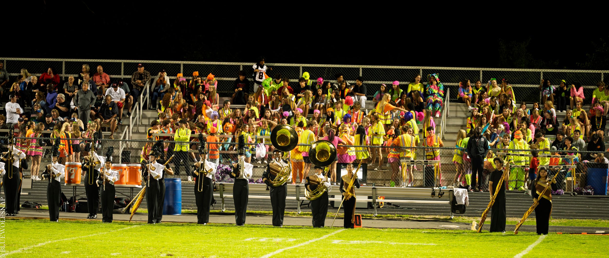 Marching band performs on football field at night with colorful uniforms and crowd in bleachers.