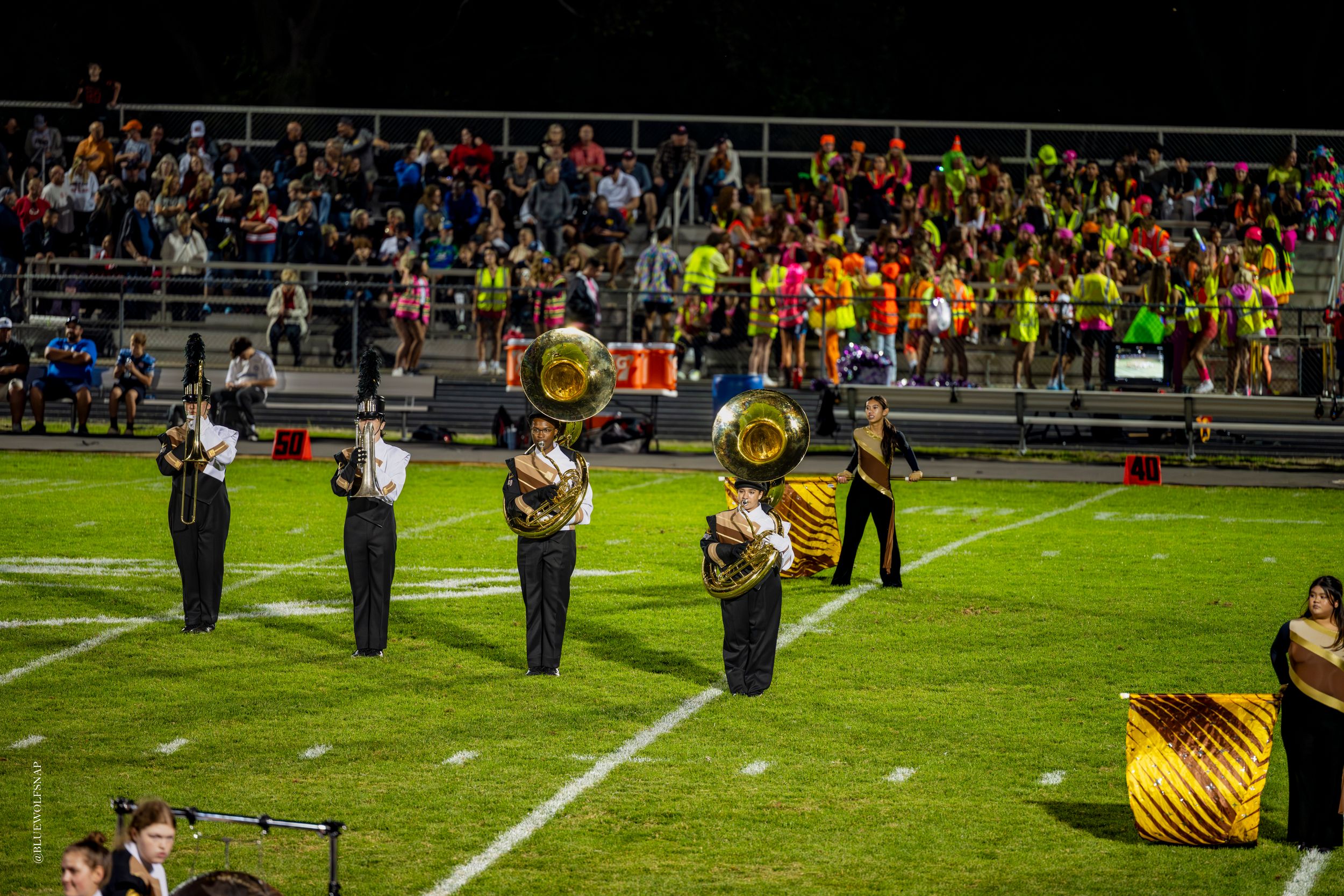 Marching band members perform on a football field at night, playing tubas in uniform before packed bleachers.