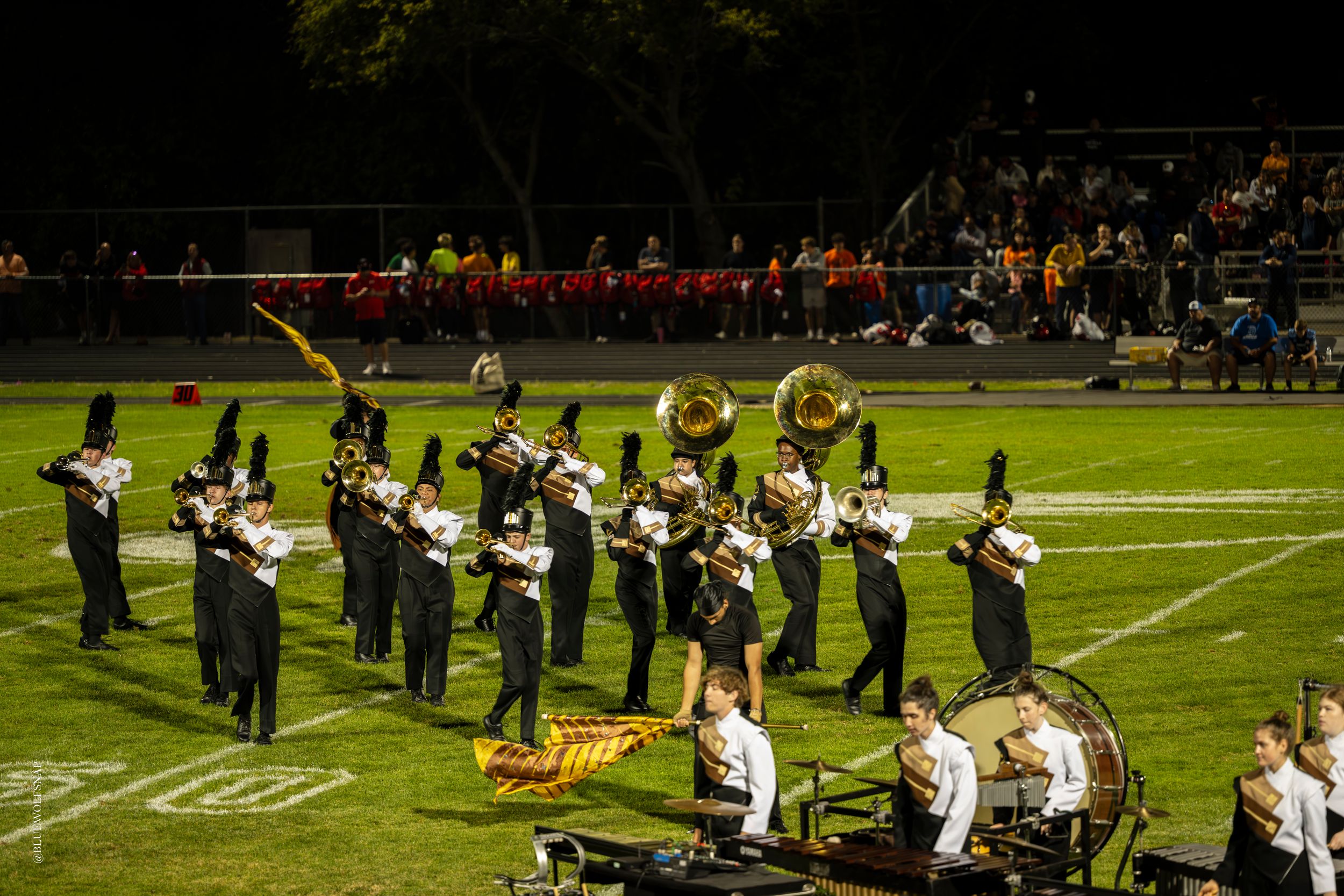 Marching band performing on a football field at night in black uniforms with brass instruments and drums.