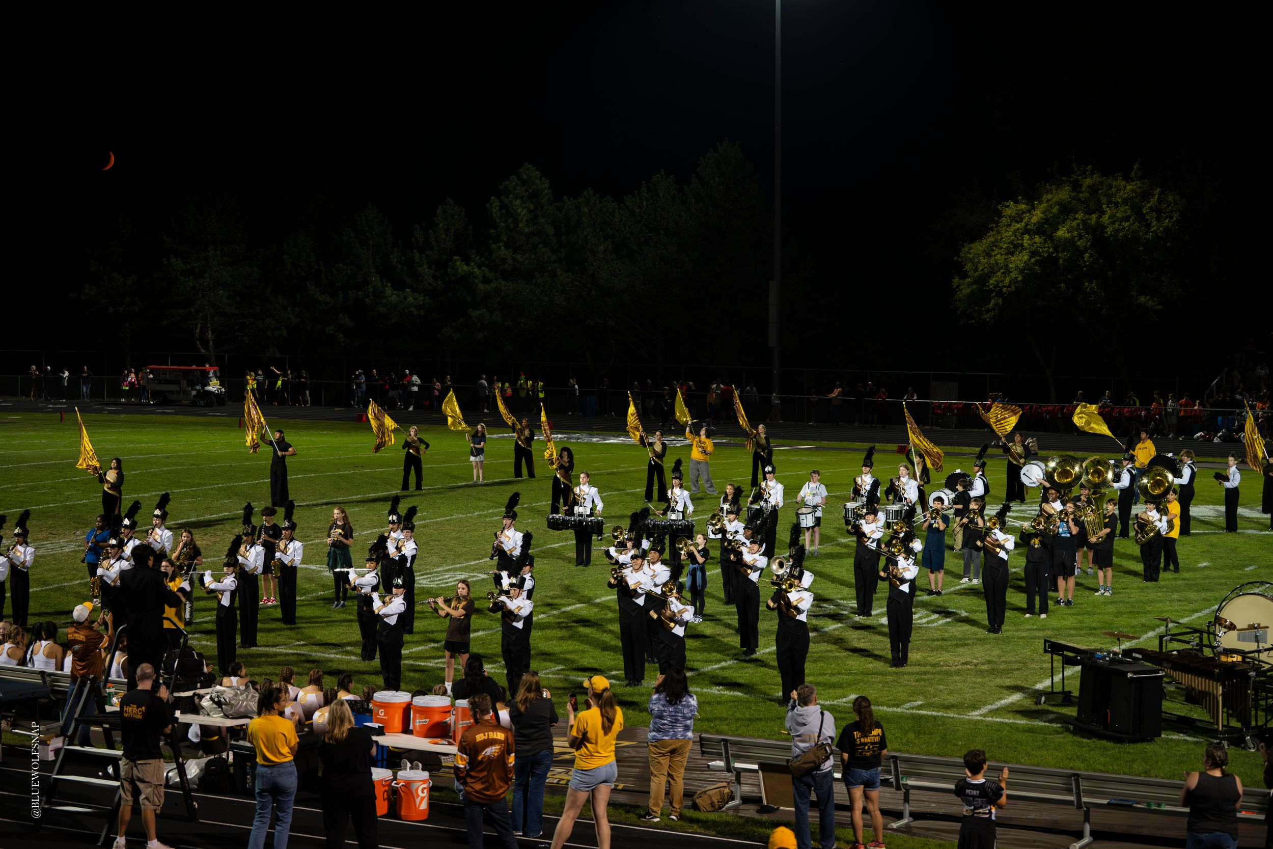 Marching band performs on football field at night with gold and black uniforms and colorful flags.