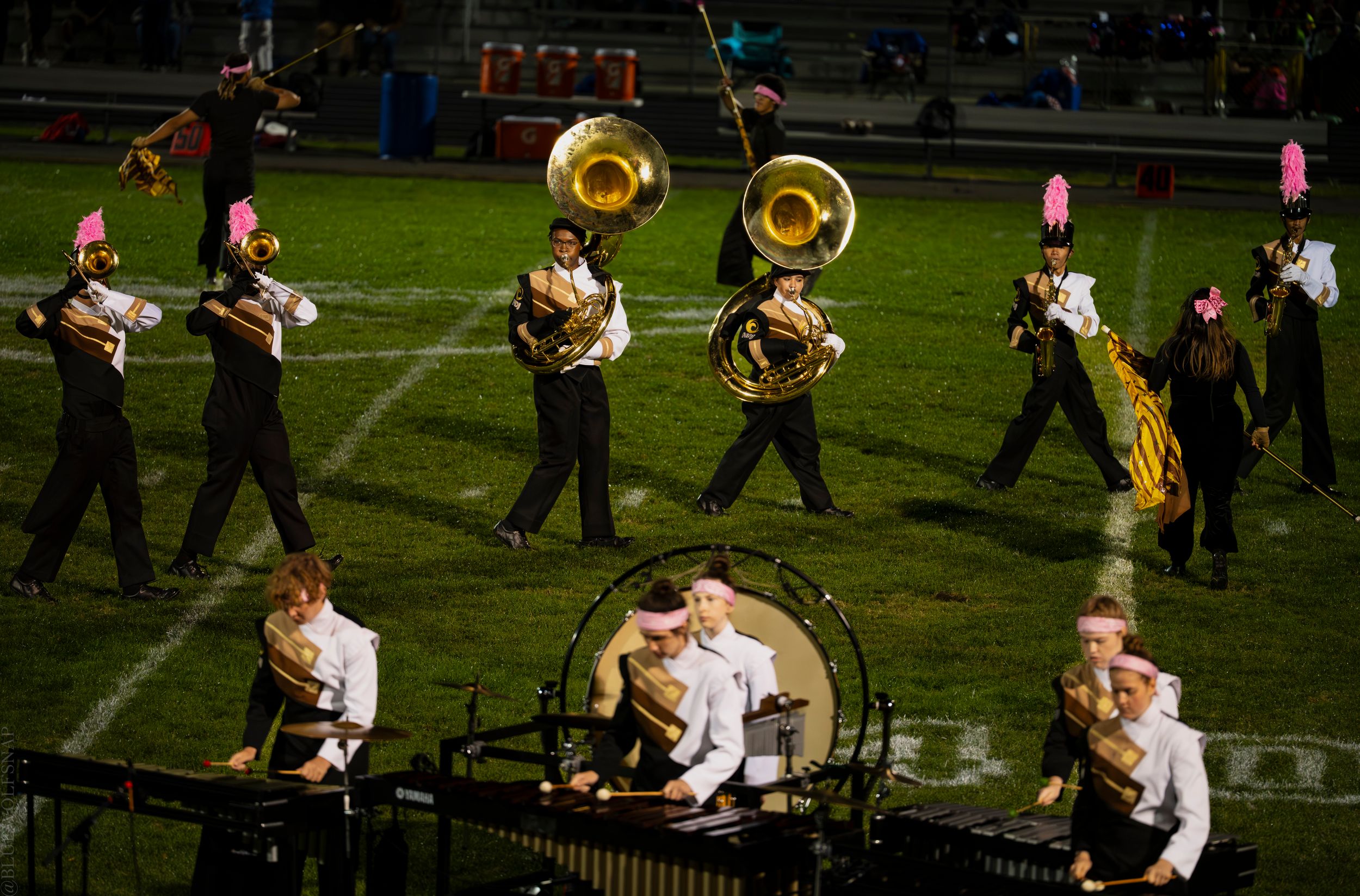 Marching band performs on football field at night, featuring tuba players and percussion section in black uniforms.
