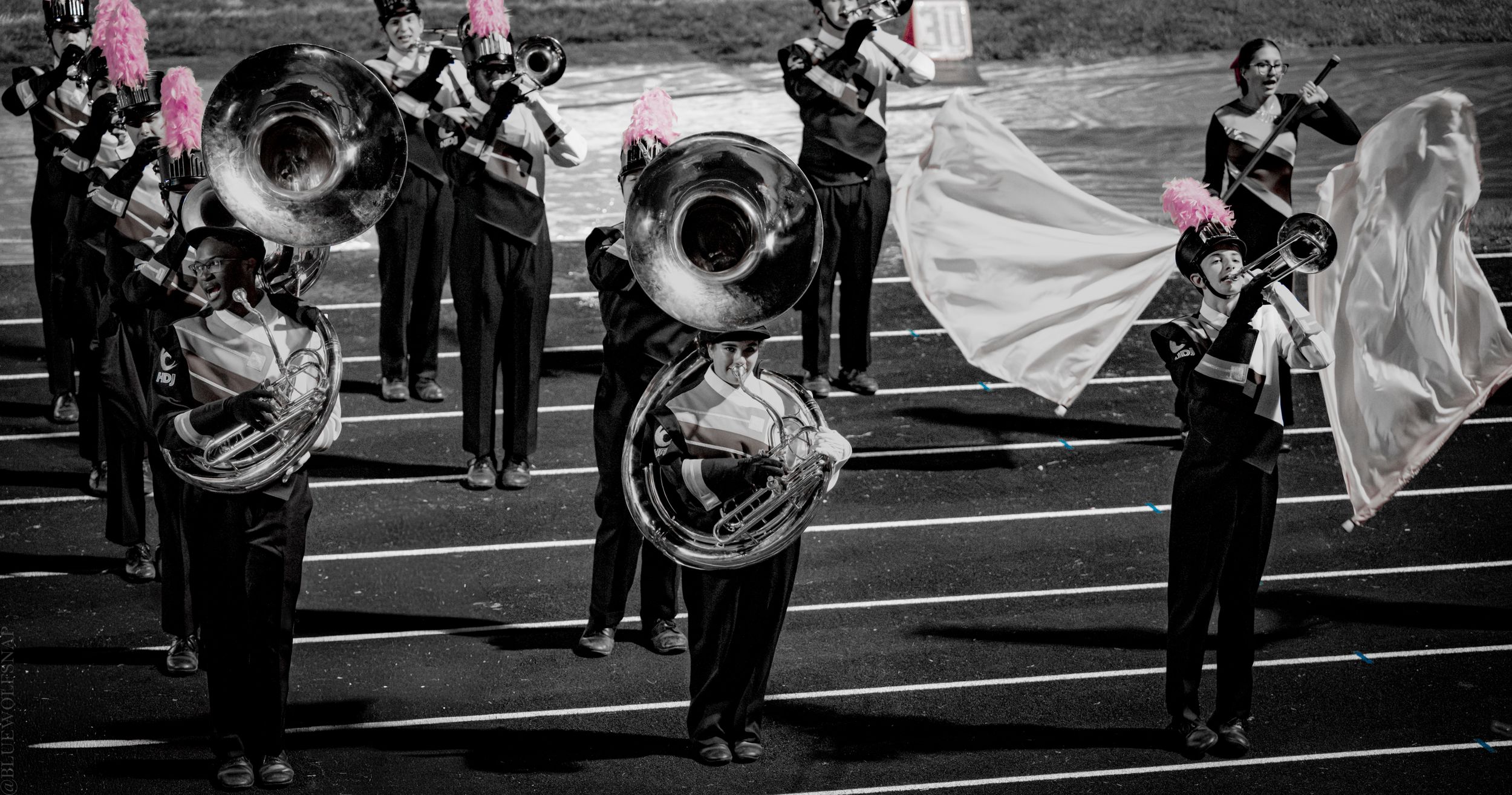 Marching band members in black uniforms play sousaphones and brass instruments on a track field during a night performance.