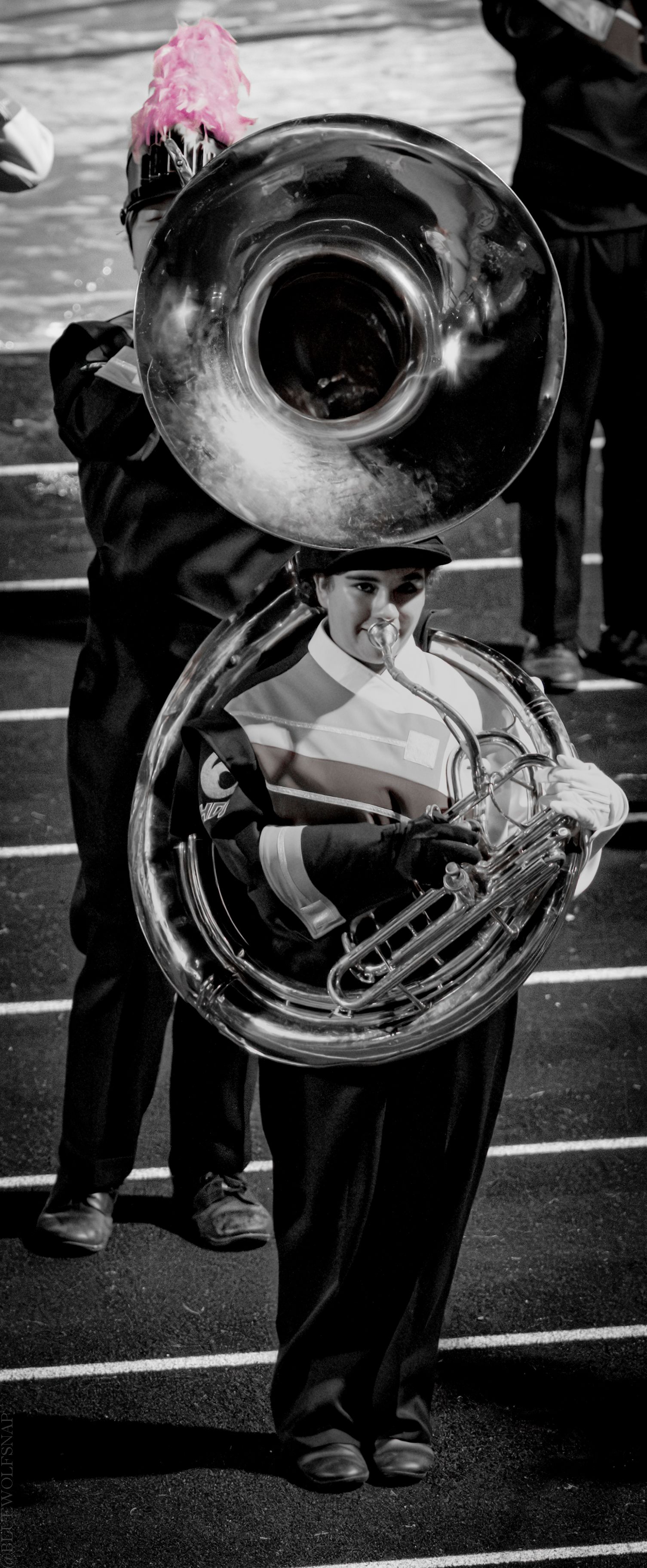 Black and white photo of a marching band member playing a sousaphone in uniform on a field.