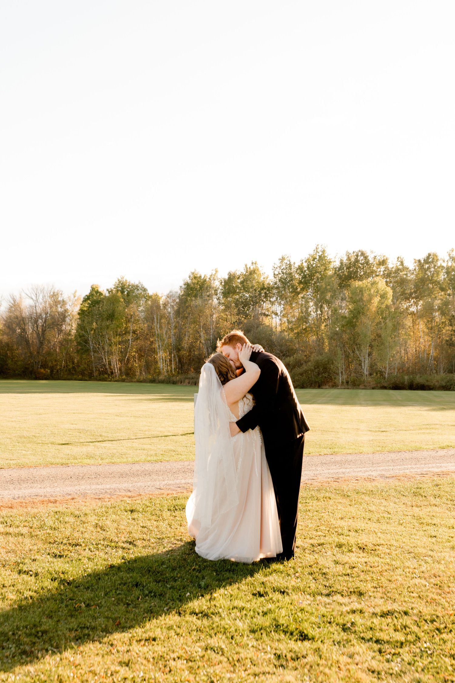 Bride and groom share a romantic embrace on a sunlit green field with autumn trees in the background.