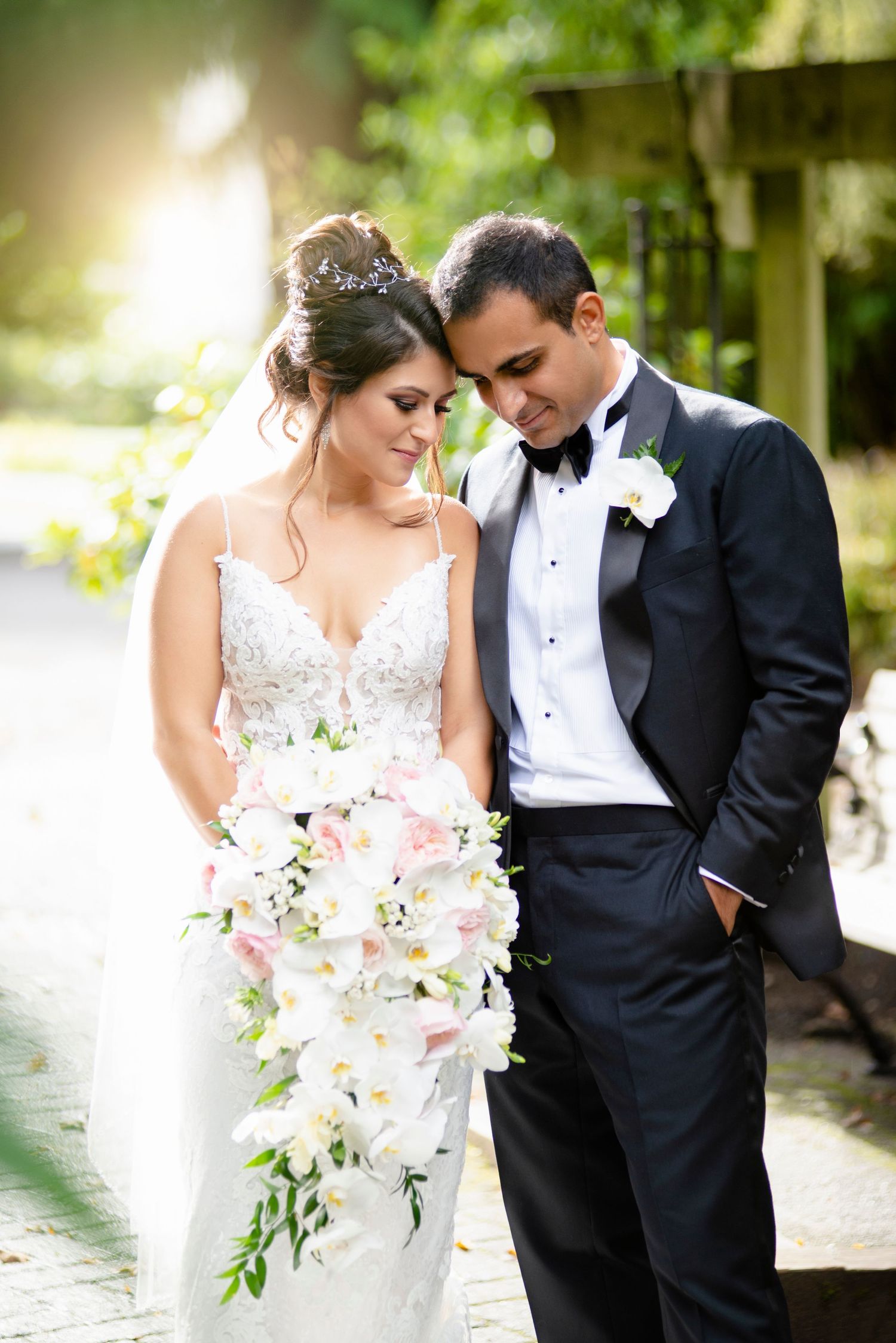 Bride holding cascading white orchid bouquet leans on groom in black tuxedo during outdoor wedding portrait.