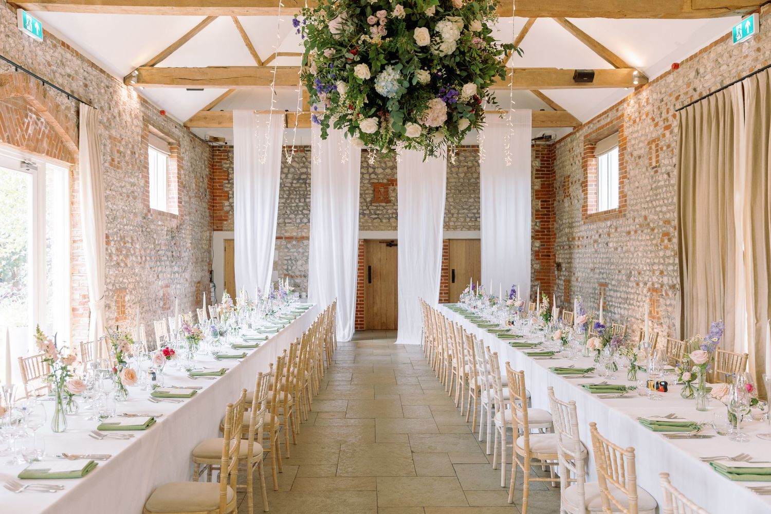 Long rustic barn venue decorated with white linens and floral arrangements for an elegant wedding reception.