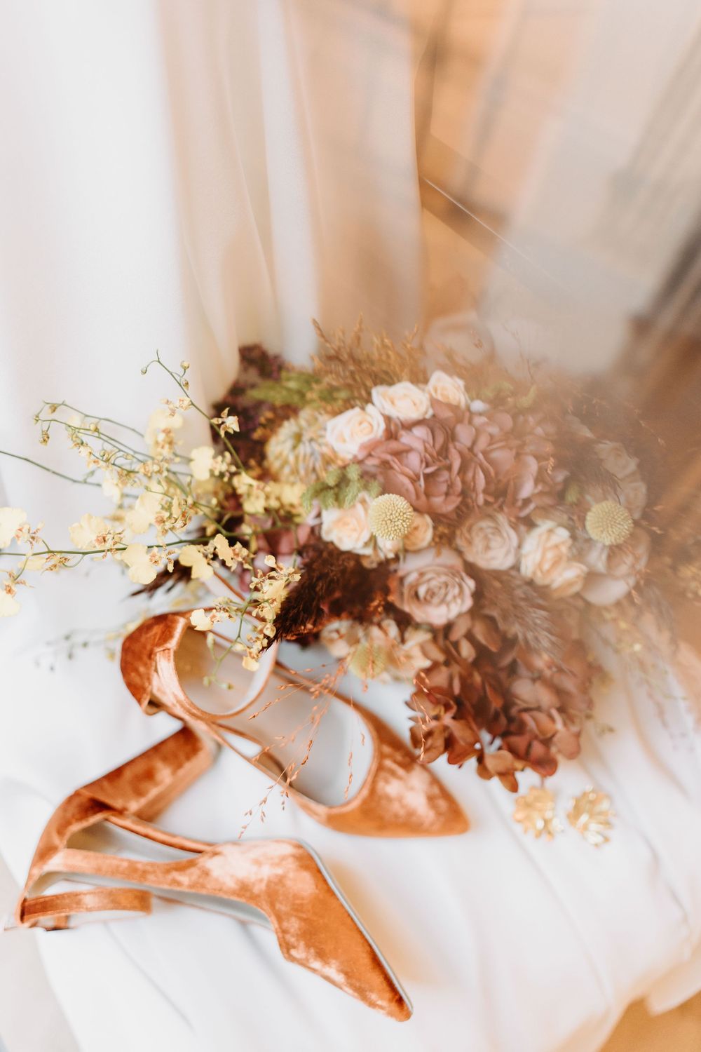 Elegant bridal bouquet of pink roses and baby's breath alongside copper-colored satin ballet flats on white fabric.