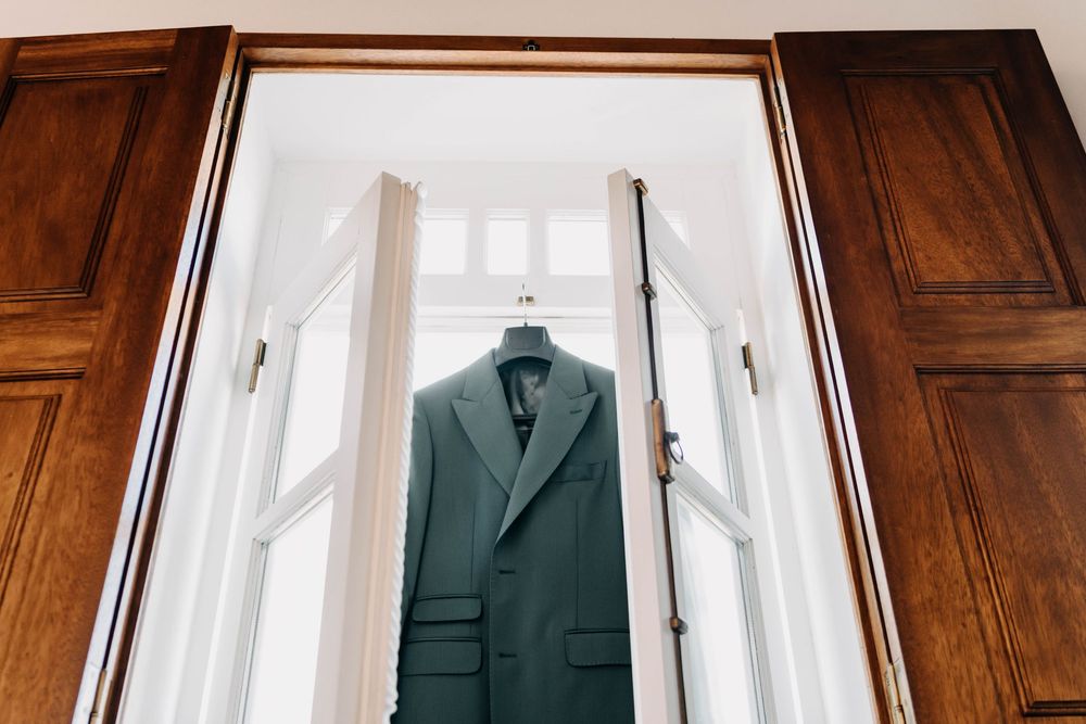 A row of green suit jackets hanging on wooden hangers against a white wall with natural lighting.