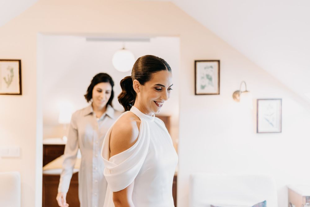 Two people in a bright white room getting ready for a special occasion, one wearing an elegant white dress.