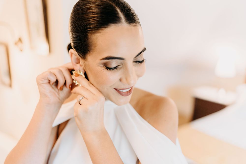 Elegant bride putting on gold earrings while smiling with soft natural makeup in a bright room.