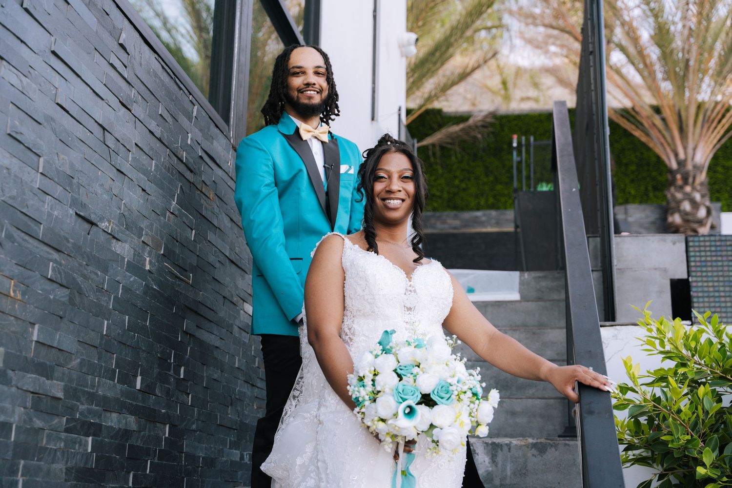 A couple in a white wedding dress and turquoise suit pose together on steps by a dark brick wall.