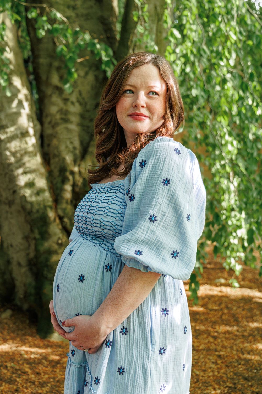 Maternity photo series showing a pregnant woman in a light blue dress posing against a backdrop of green trees.
