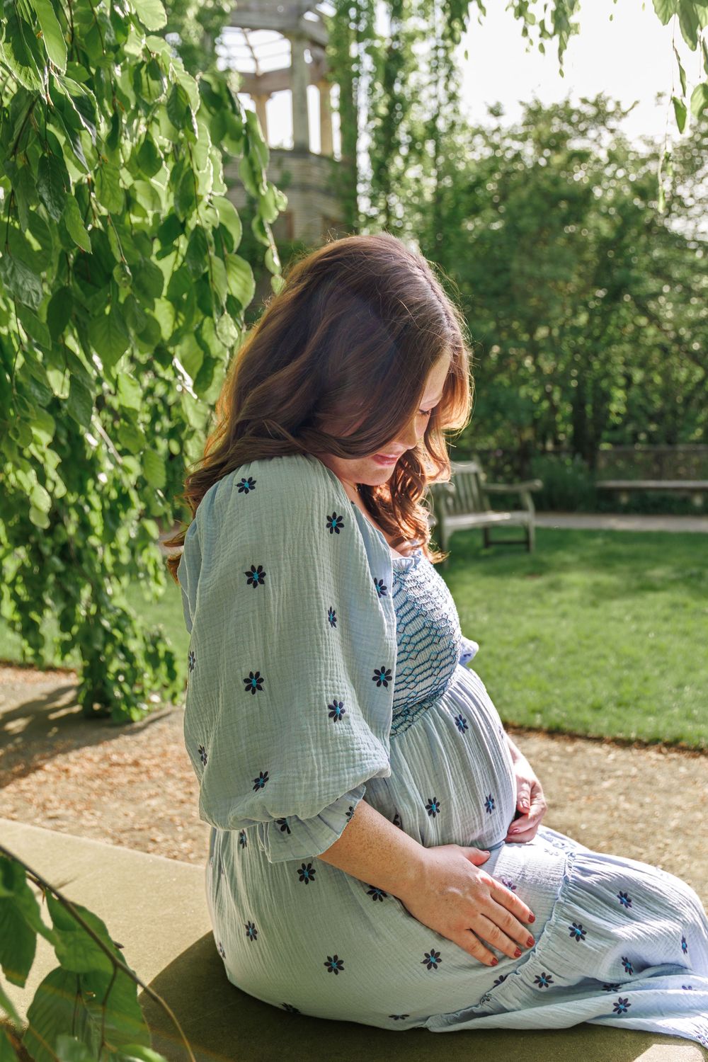 A pregnant woman in a floral dress sits peacefully in a garden setting with green foliage surrounding her.