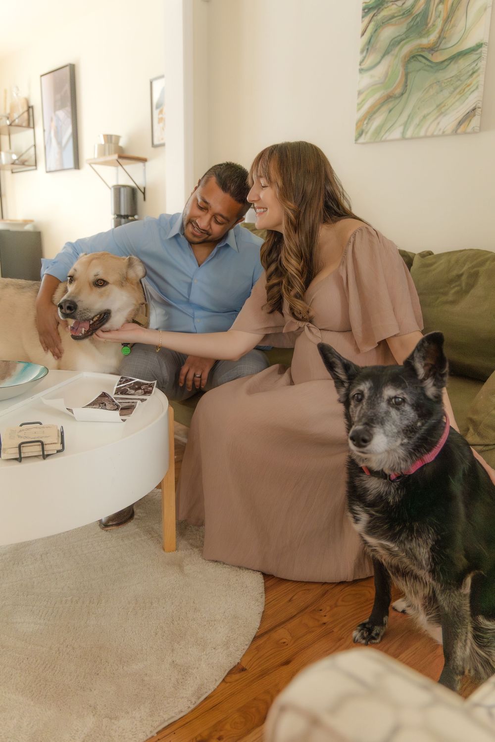 A couple sits on a couch looking at photos while their senior dog rests nearby on the hardwood floor.