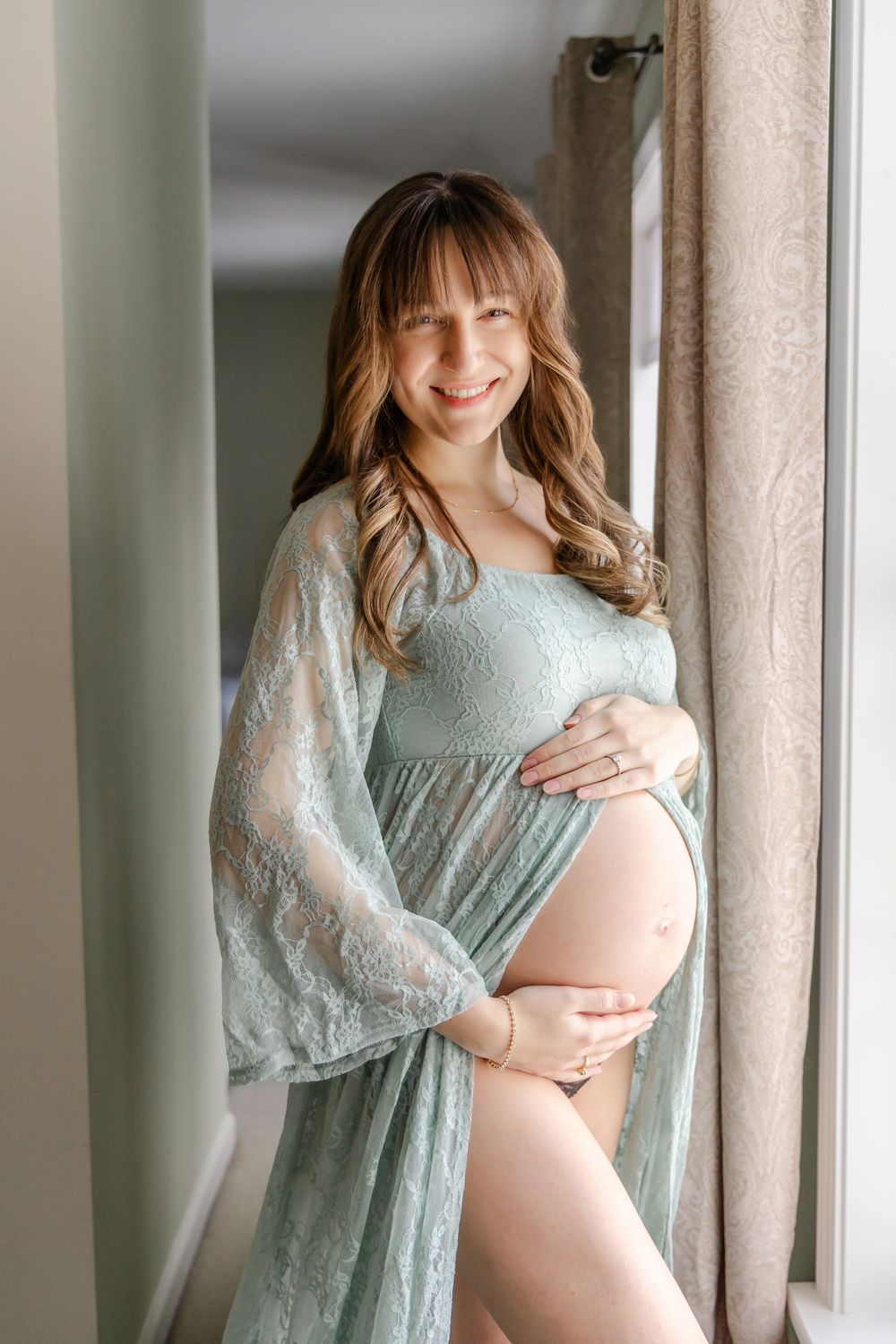 A maternity photoshoot featuring a woman in a light blue lace dress posing by a window.