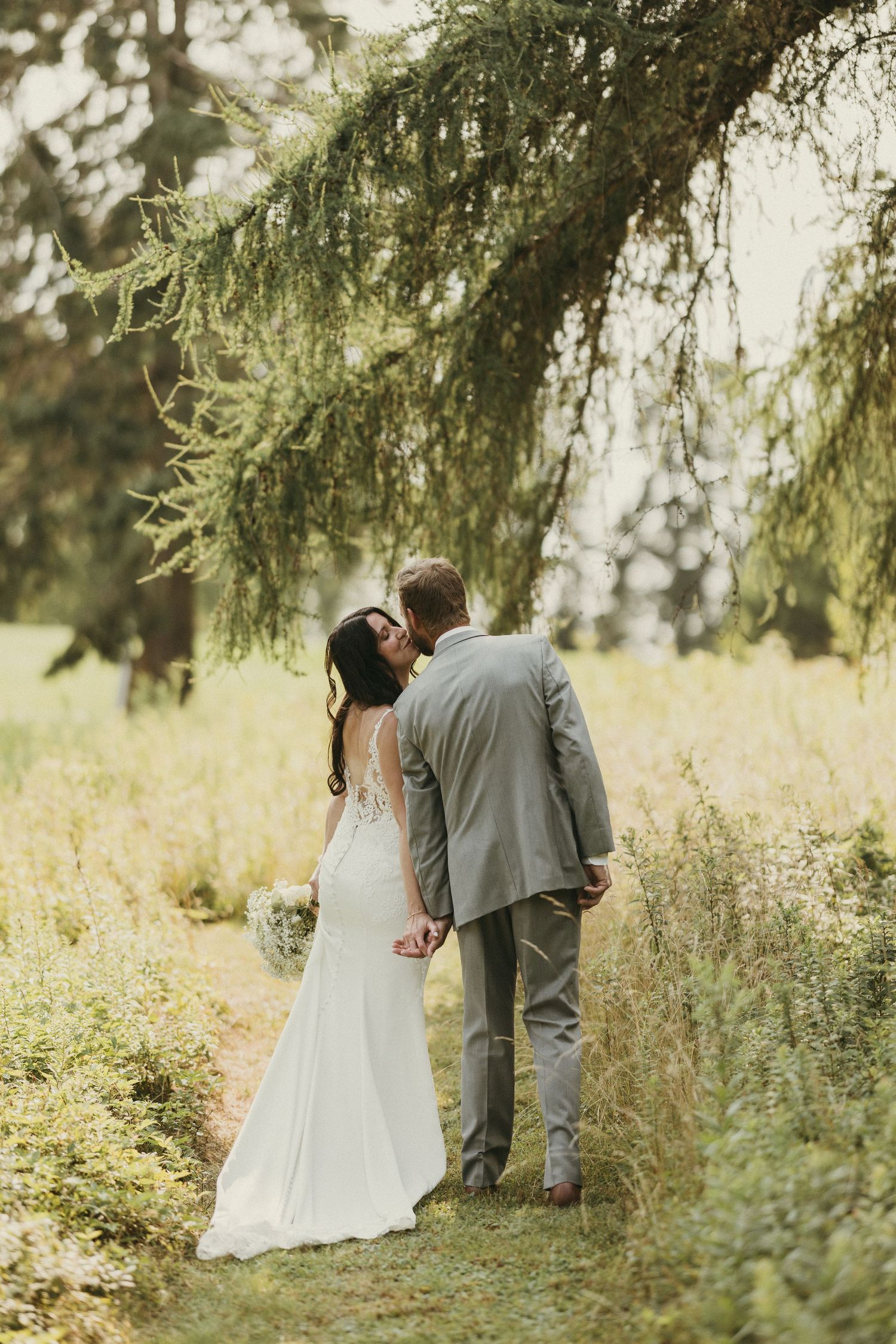 A romantic wedding couple shares intimate moments under overhanging trees in a dreamy outdoor setting.