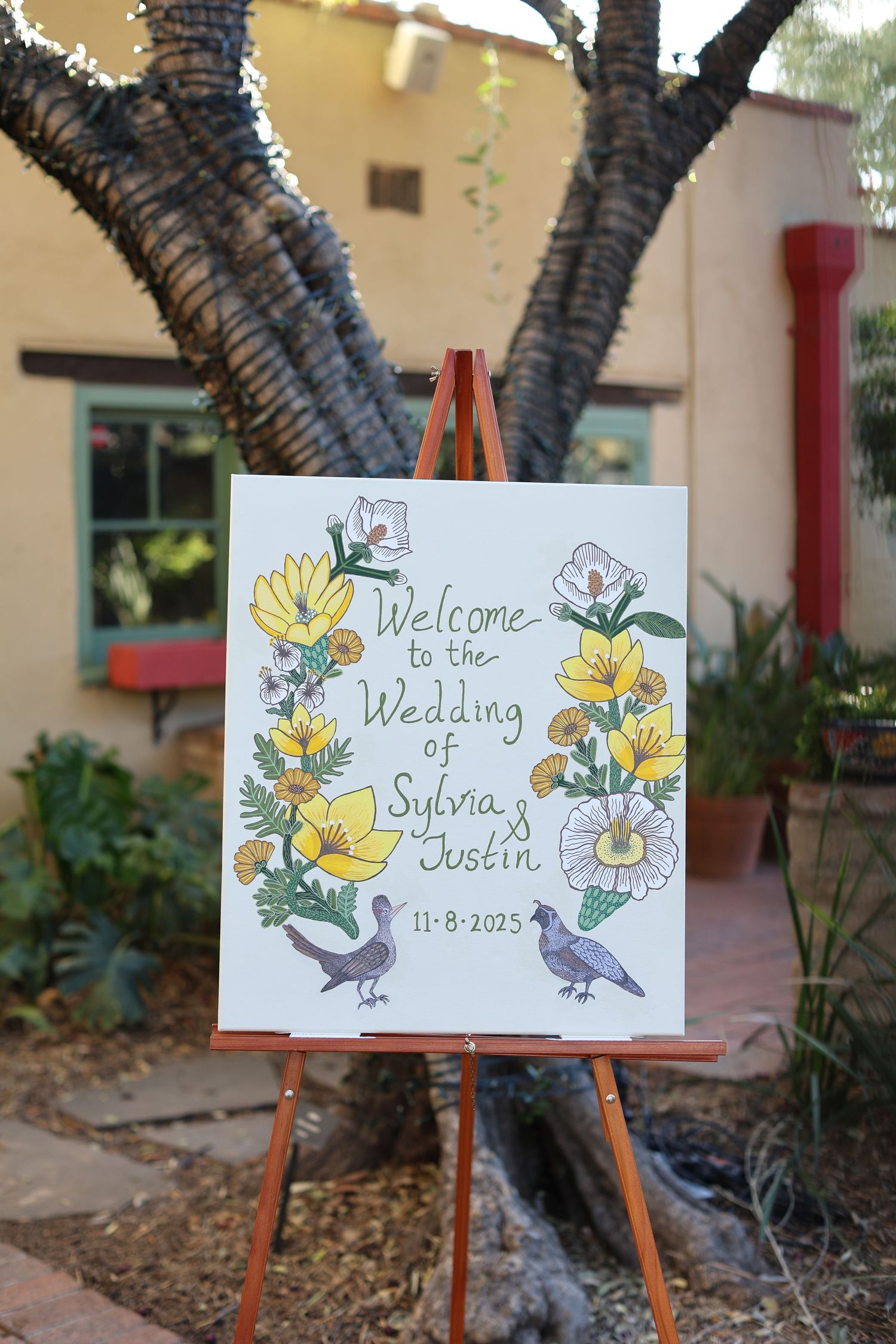 Hand-drawn wedding welcome signs with yellow flowers and birds displayed on wooden easels in an outdoor garden setting.