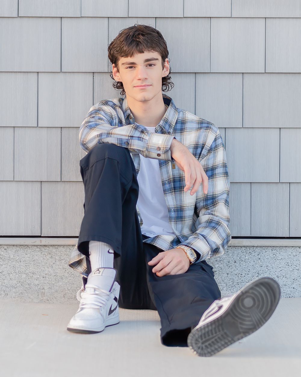 A high school senior in a plaid shirt and dark pants sitting against a gray wall in Bedford, NH