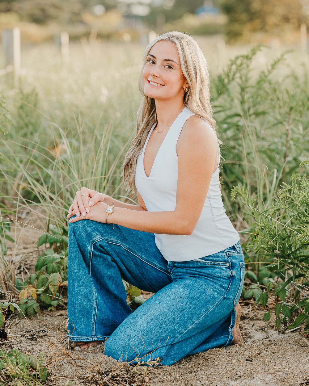 A high school senior in a white sleeveless top and blue jeans sits casually outdoors in a grassy field during golden hour in Plum Island, Ma