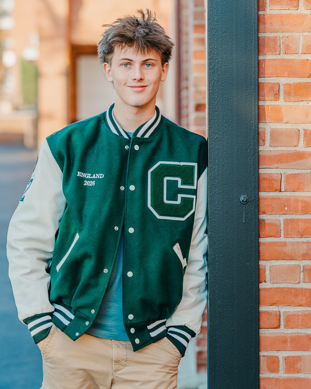 A male high school senior in varsity letterman jacket shown from different angles against a brick wall background in Manchester, NH