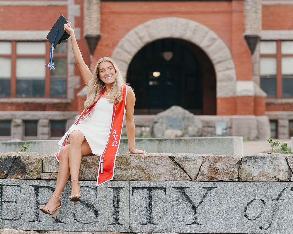 College graduate in white dress sits on stone wall with orange scarf blowing in wind against brick building backdrop at UNH in Durham, NH