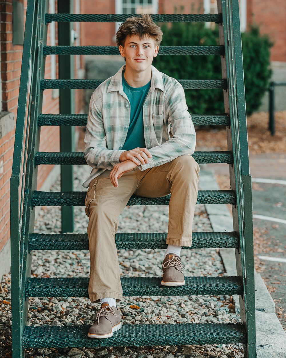 A male high school senior in khaki pants and light jacket sits on green metal stairs in an urban setting in Manchester, NH