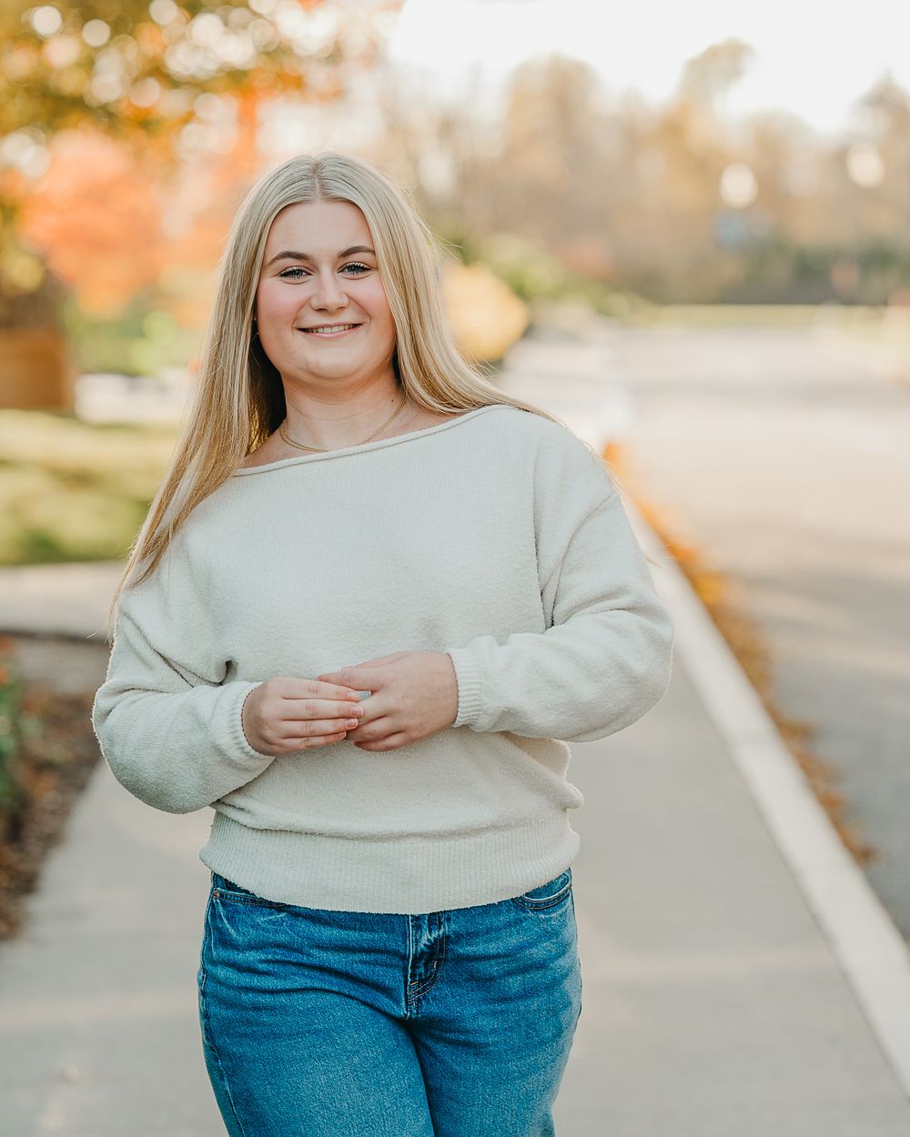 A female high school senior  in cream sweater and blue jeans poses outdoors during golden hour with autumn scenery in Goffstown, NH