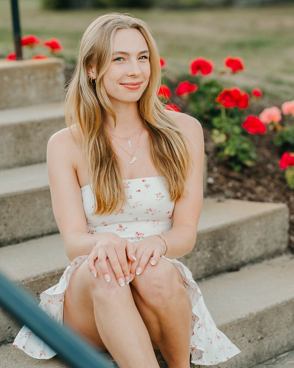 High school senior in a white floral dress sits on concrete steps near blooming red flowers in a garden setting in Goffstown, NH