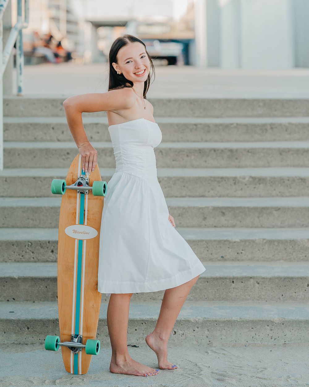 High school senior in white sundress poses playfully with turquoise longboard skateboard on concrete steps in summer in Hampton Beach, NH