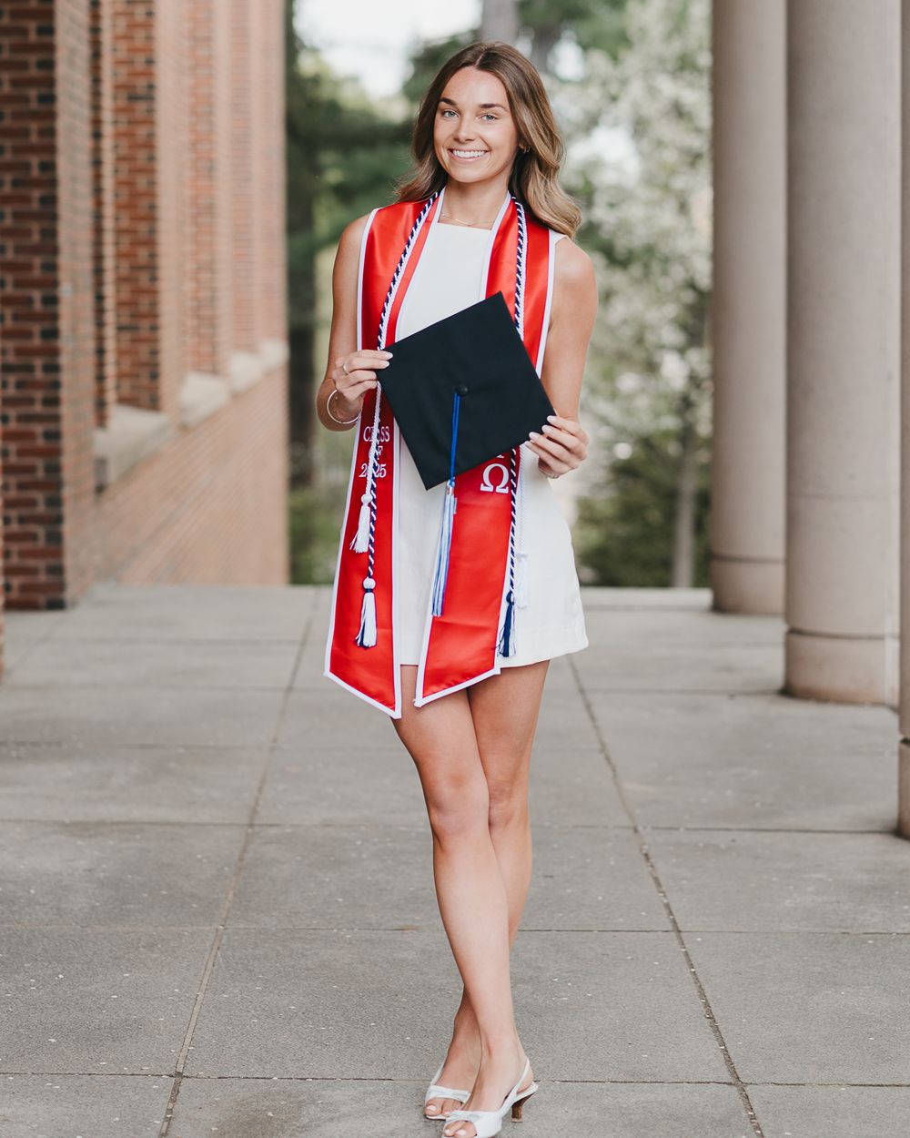 College Graduate in white dress and red graduation stole poses outdoors near brick building columns at UNH in Durham, NH