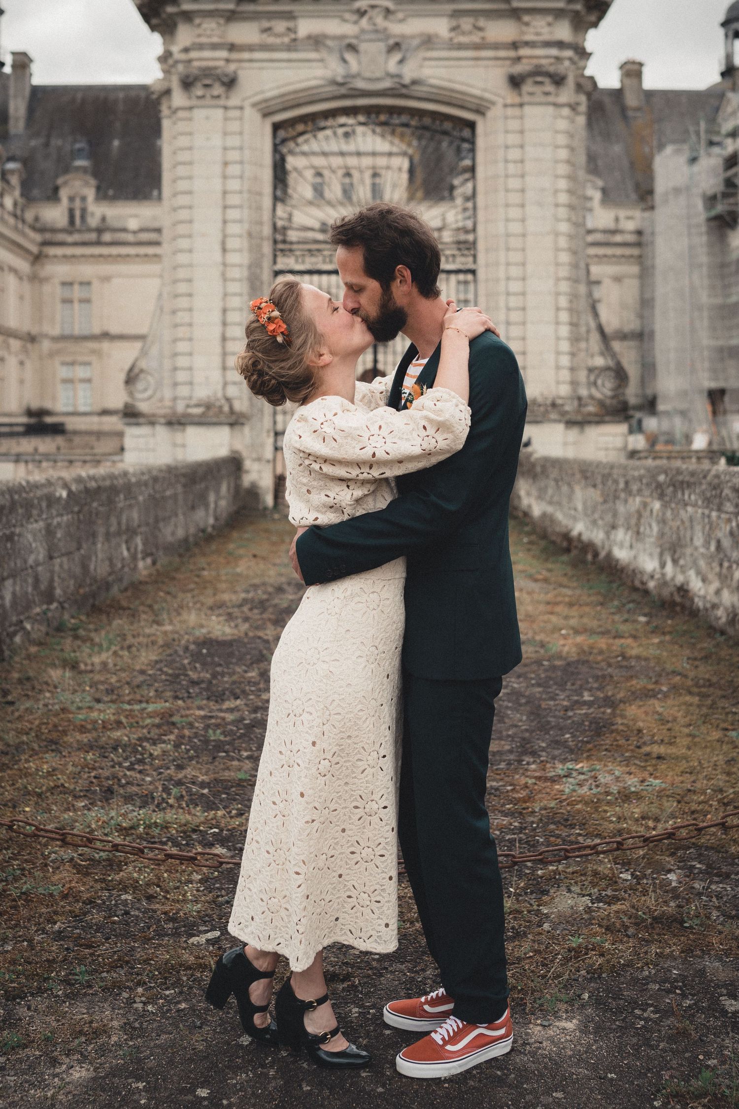 A romantic couple embraces on a historic castle grounds in winter, with ornate architecture visible in the background.