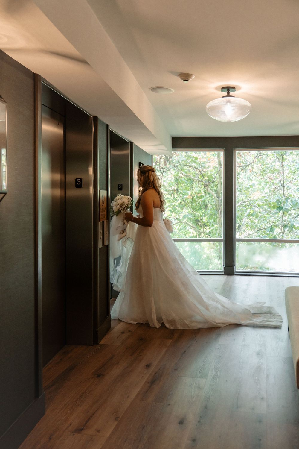 A bride in a flowing white wedding gown with a long train walks down a modern hallway with wooden floors and windows.