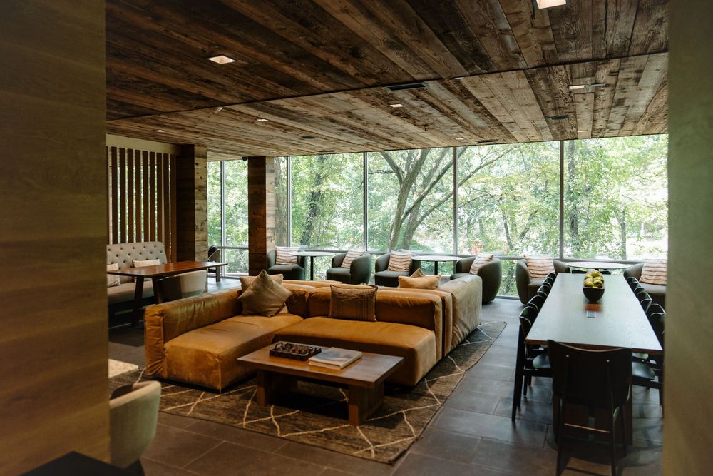 Modern living room with rustic wood ceiling, leather sofas, and large windows overlooking a wooded landscape.