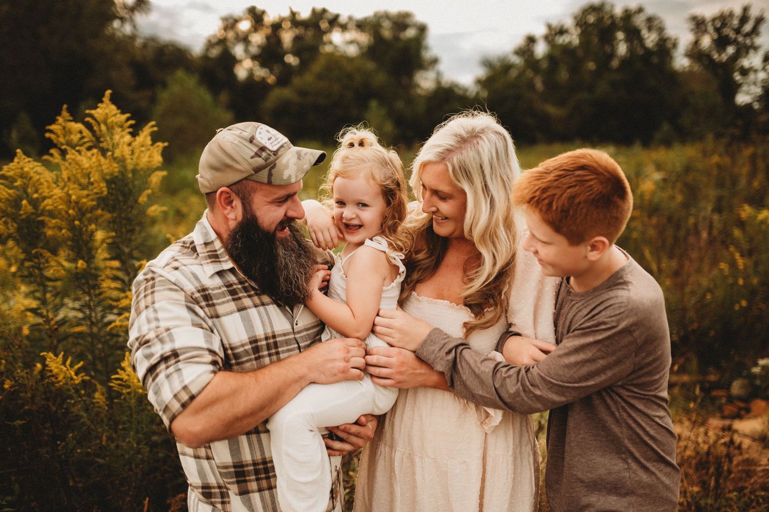 A family wearing coordinated neutral outfits embraces in a golden autumn field during sunset.