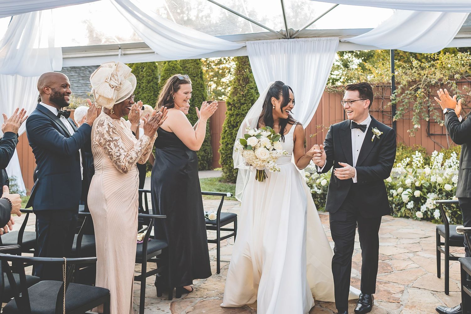 Wedding ceremony guests clap and celebrate during an outdoor celebration under a white tent with floral decorations.