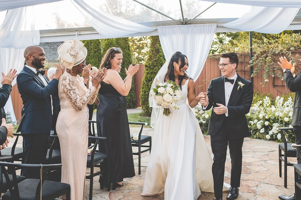 Wedding ceremony guests clap and celebrate during an outdoor celebration under a white tent with floral decorations.