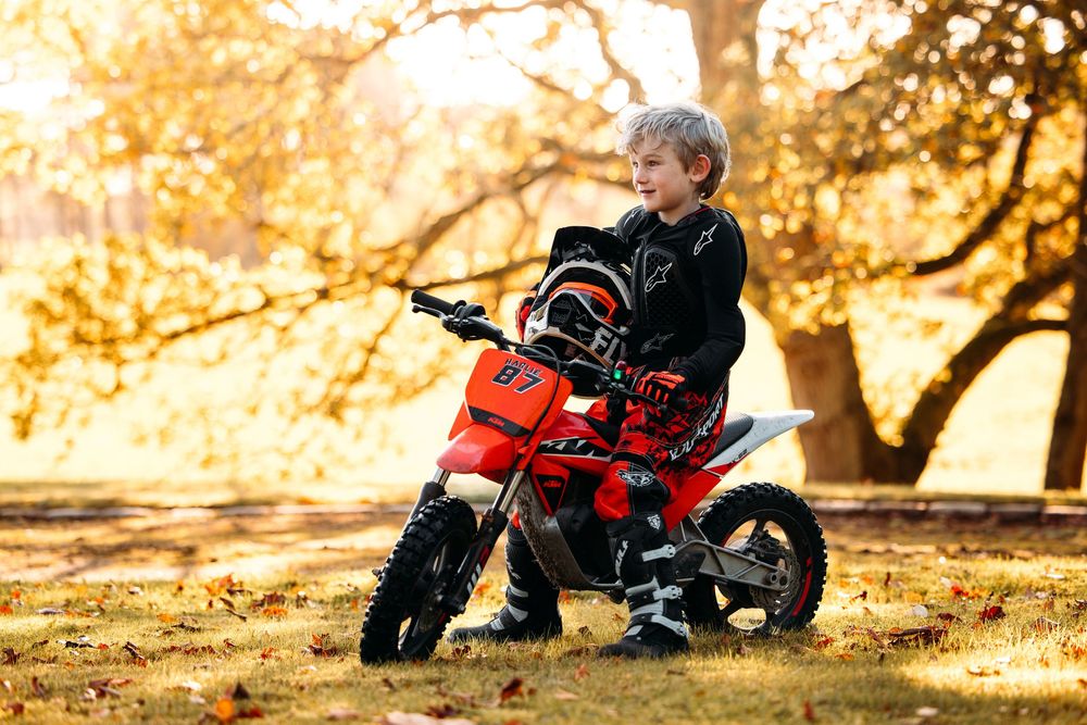 Young child riding a red mini dirt bike in golden autumn sunlight.