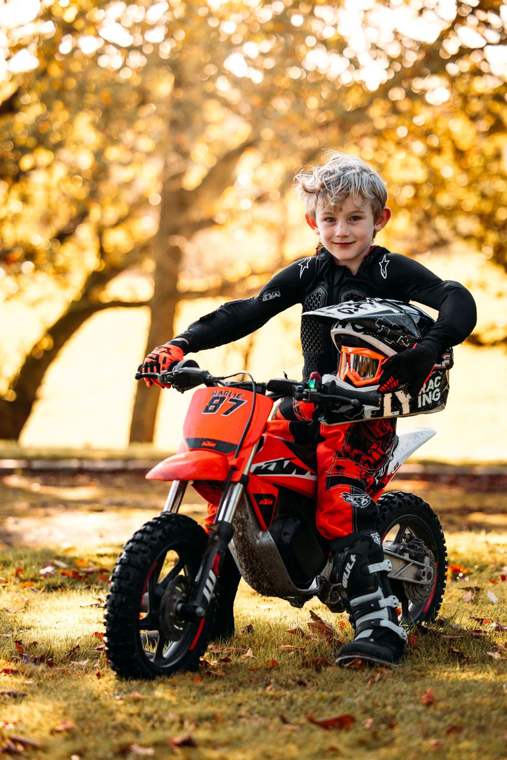 Small dirt bike rider poses with orange motorcycle against golden autumn backdrop.