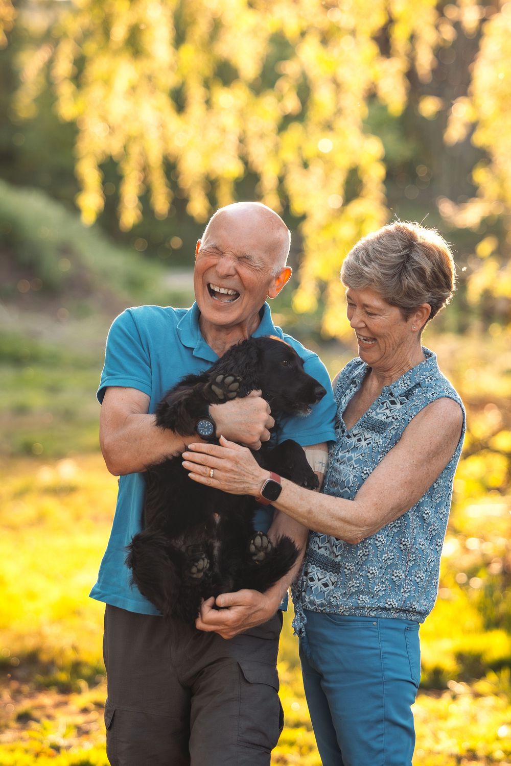 A couple shares joyful moments with their black dog in a golden-lit outdoor setting.
