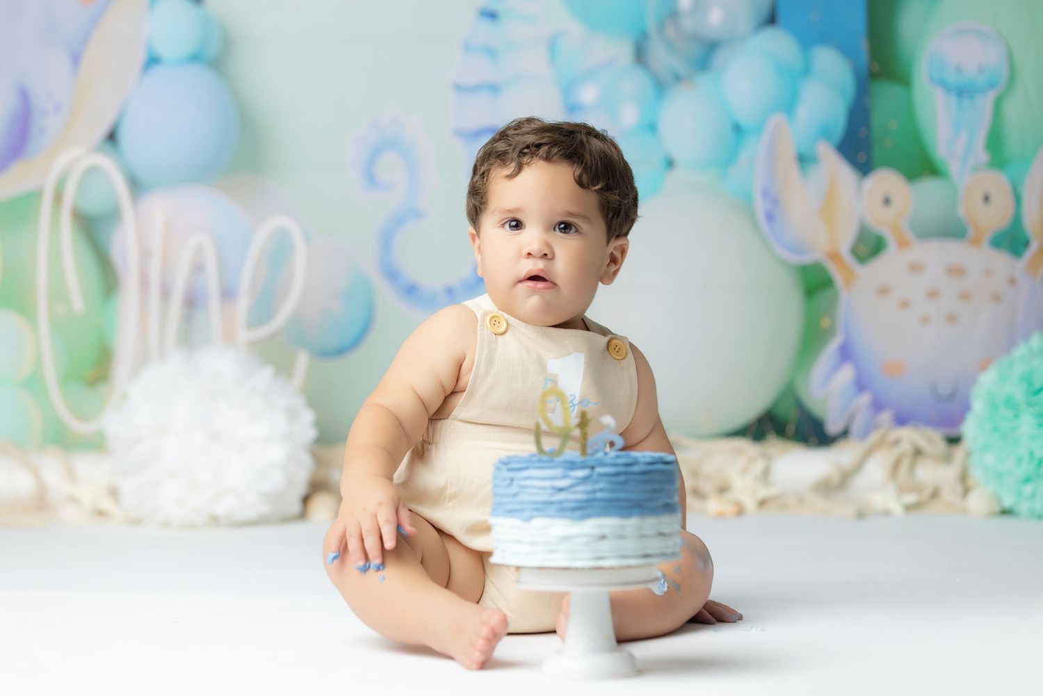 A baby wearing a lace dress sits with a birthday cake during a cake smash photo session.