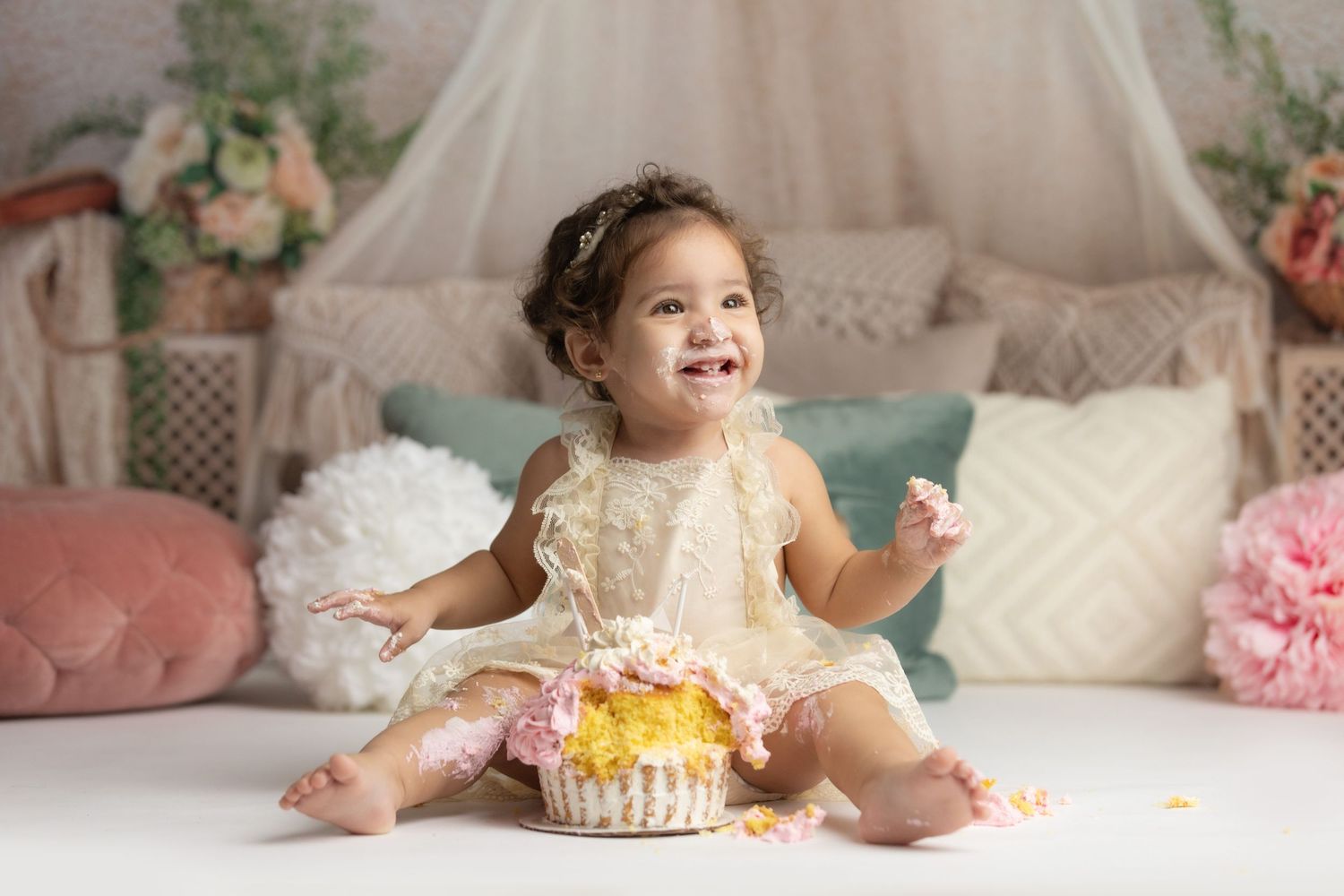 A baby wearing a lace dress sits with a birthday cake during a cake smash photo session.