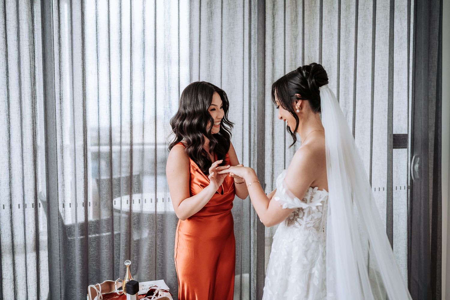Two women share an intimate moment as bride in white gown and guest in coral dress hold hands during wedding preparations.