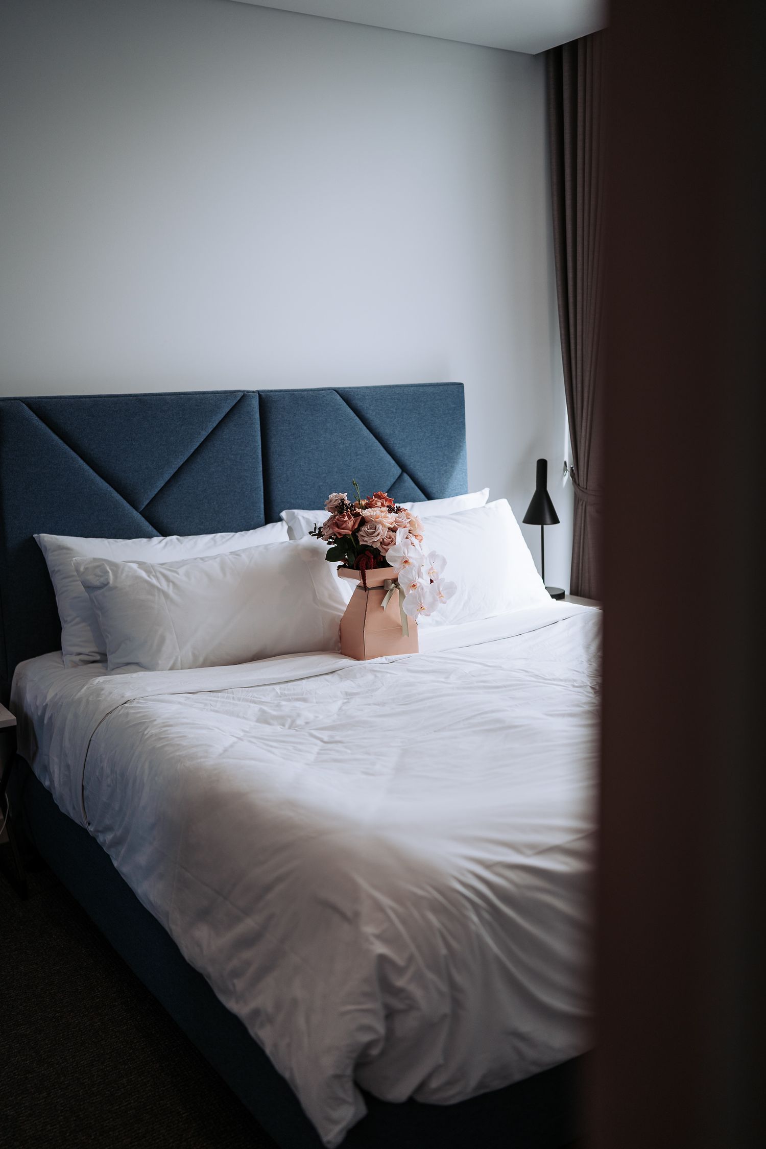 Modern bedroom with navy blue upholstered headboard, white bedding, and pink flowers in a terracotta vase on the bed.