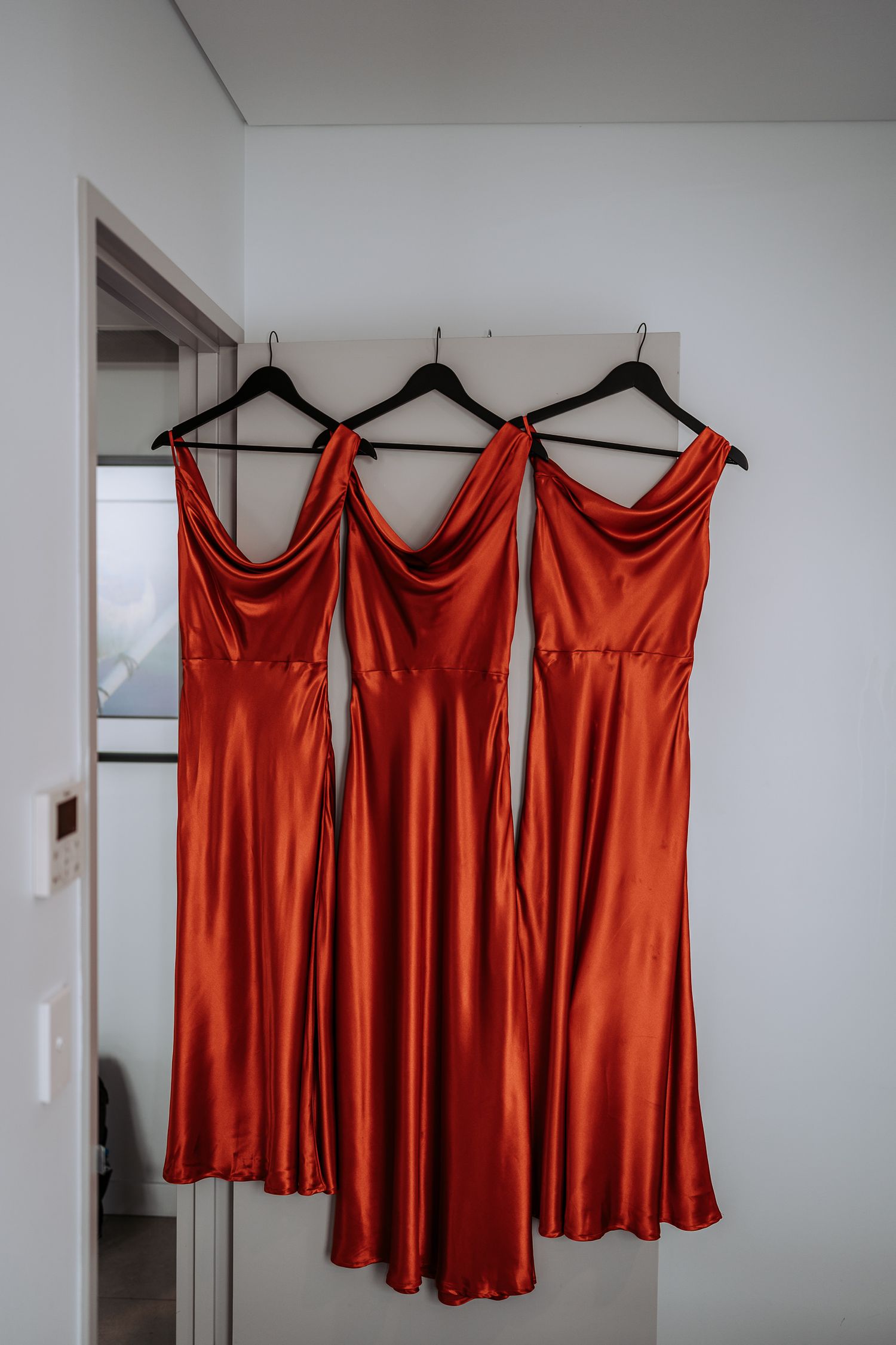 Three elegant burnt orange satin bridesmaid dresses hanging on black hangers against a white wall.